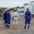 A group of construction workers, wearing blue and white protective gear with yellow and red helmets, stand on a dirt construction site. Portable buildings are visible in the background along with construction equipment.