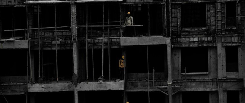 A construction site featuring a multi-story building in progress. Several workers wearing yellow helmets are scattered across different floors, with some engaged in activities such as carrying materials or working on the structure. The building appears to be made of concrete, and there are numerous steel rods protruding from the top floor.
